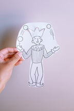 Hand holding paper of a jester with a crown and juggling balls against a light gray background
