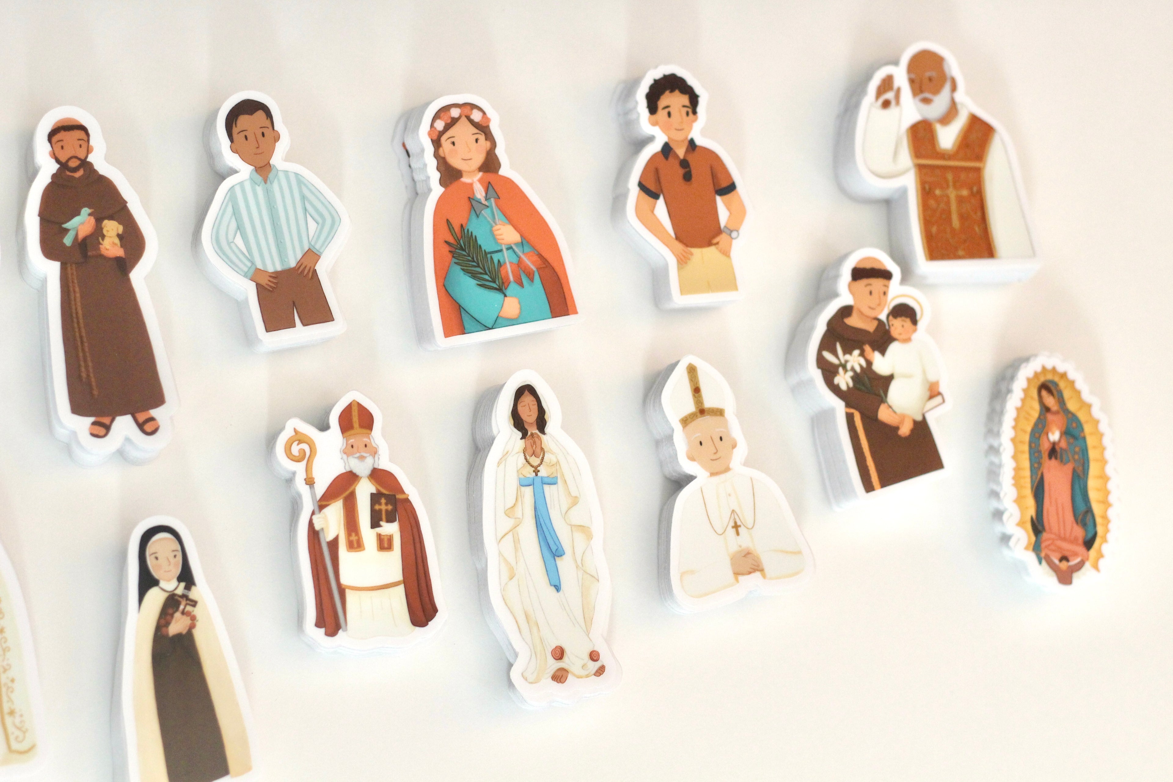 A set of small illustrated religious figurines or stickers shown on a plain white background. The figures depict various saints, priests, and religious icons in simple cartoon-style drawings with white borders around each character.
