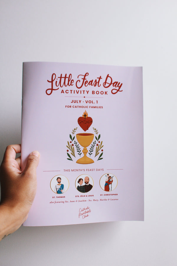 Hand holding a 'Little Feast Day Activity Book' on a white background