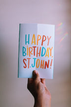 Hand holding a colorful 'Happy Birthday, St. John!' card against a plain background