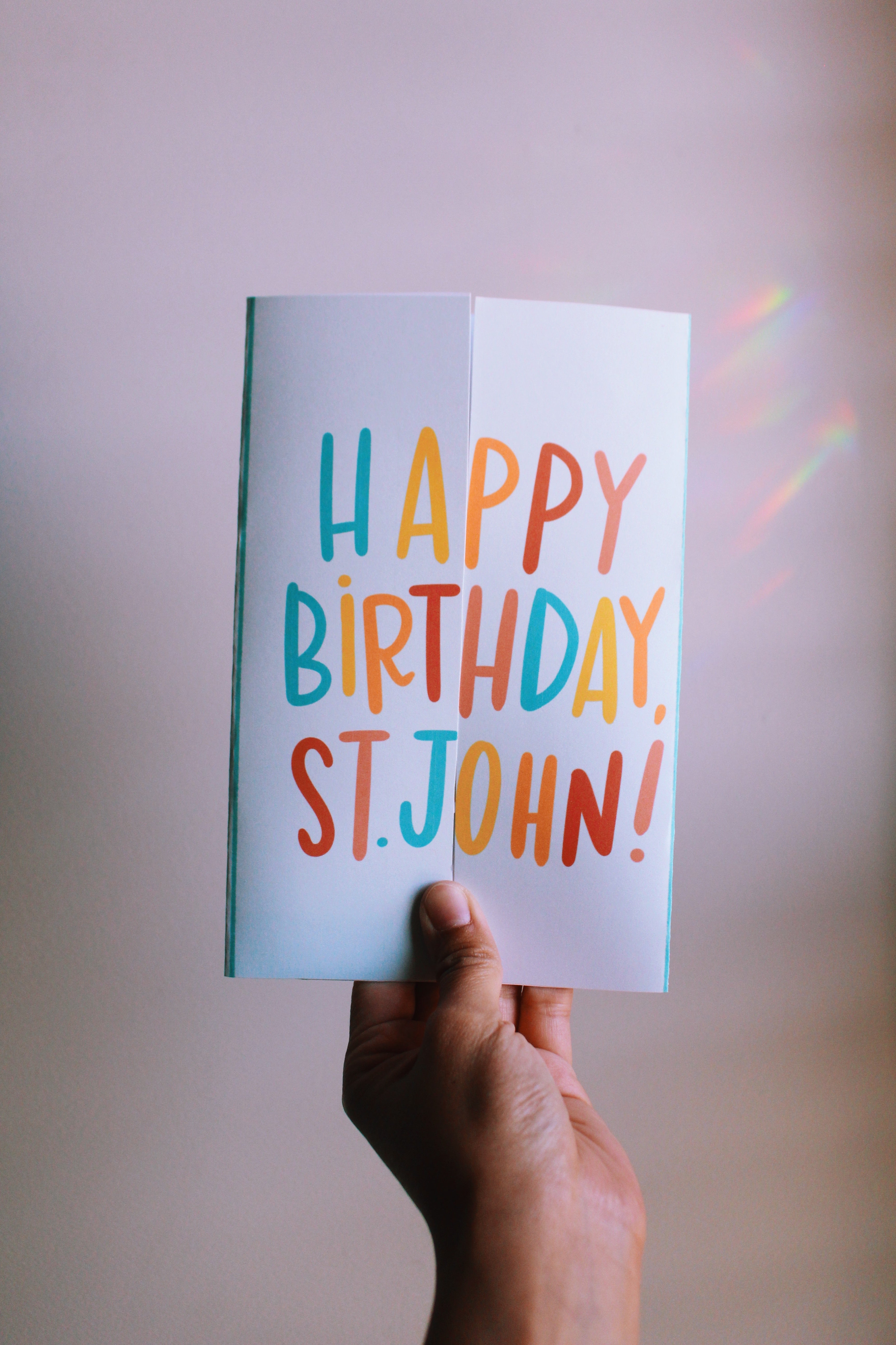 Hand holding a colorful 'Happy Birthday, St. John!' card against a plain background