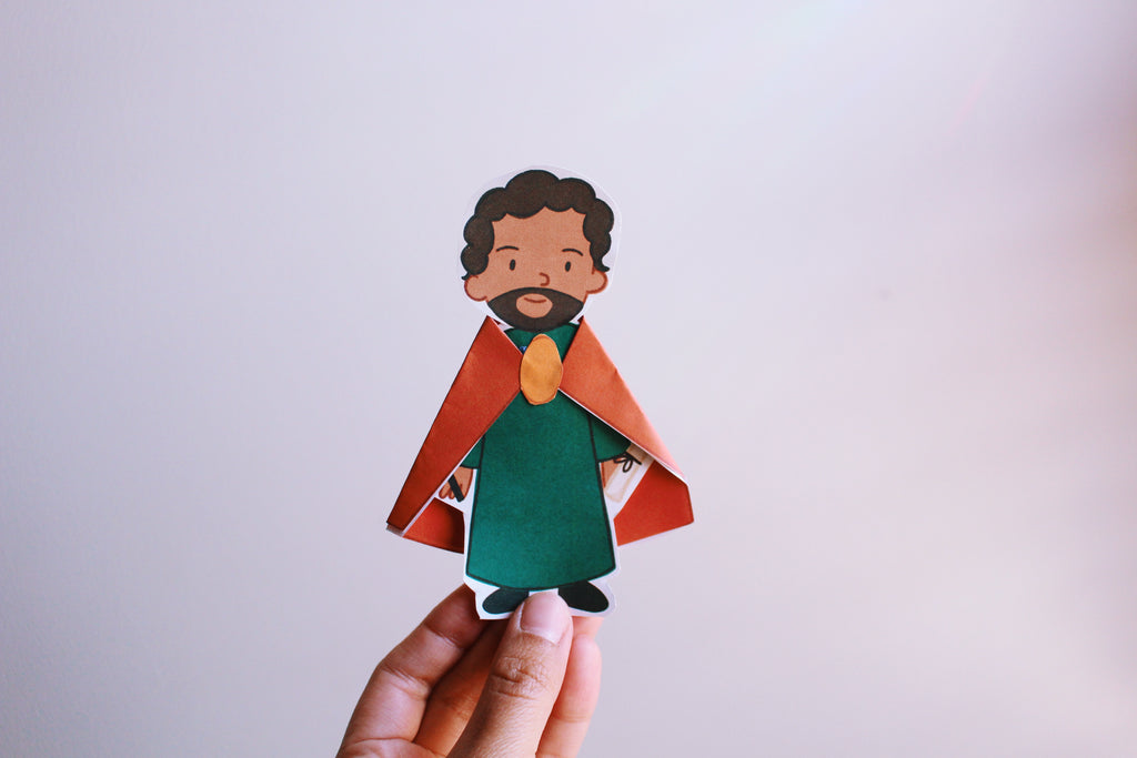 Hand holding a paper cutout of a character with a red cape and green outfit against a light background